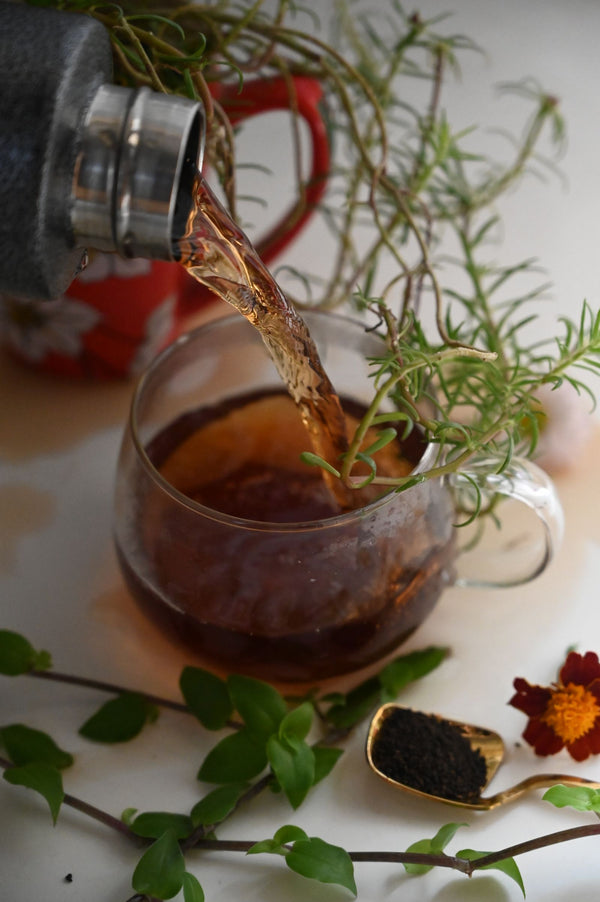 Herbal tea being poured into a glass cup surrounded by fresh green herbs