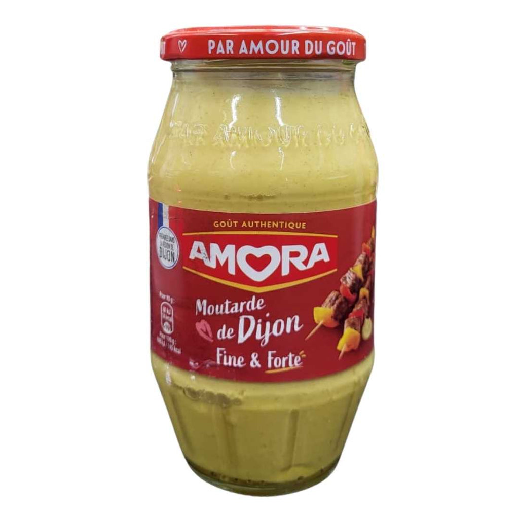 Jar of Amora Dijon Mustard (Moutarde de Dijon Fine & Forte) with red label and cap, displayed on a store counter with shelves of grocery items in the background.